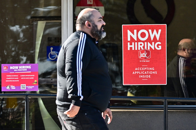 A pedestrian walks past a "Now Hiring" sign posted on a business storefront